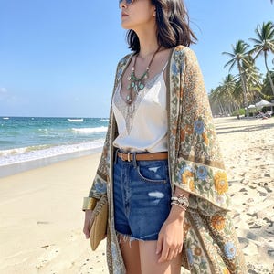 May include: A beige kimono featuring a floral pattern in orange and blue, worn over a white camisole with lace trim and blue denim shorts. The outfit includes a tan leather belt, a beaded necklace with turquoise accents, and a gold-tone cuff bracelet.