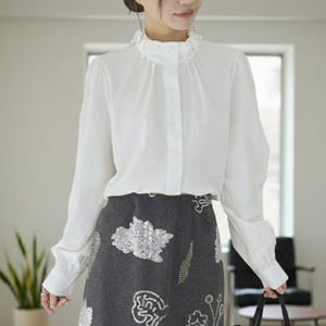 Pode incluir: Uma blusa branca de manga comprida com gola com babados e fecho de botões. A blusa é combinada com uma saia cinza adornada com lantejoulas prateadas. O conjunto é elegante.