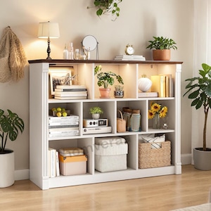 May include: A white wooden bookshelf with a dark brown top, featuring multiple compartments filled with books, plants, decorative items, and storage boxes. The shelf is illuminated with built-in lighting, creating a warm and inviting display.