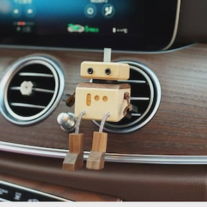 Könnte beinhalten: Ein hölzerner Roboter-Clip für die Lüftung im Auto. Der Roboter hat einen hellfarbigen Kopf und Körper mit dunkelbraunen Akzenten. Er hat kleine schwarze Augen und Holzbeine. Der Roboter ist an einer Lüftungsöffnung im Auto befestigt.