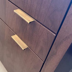May include: A dark brown wooden cabinet featuring two stacked drawers. Each drawer is equipped with a horizontal, brushed brass-toned metal pull handle that is mounted along the top edge of the drawer front, providing a sleek and functional grip.