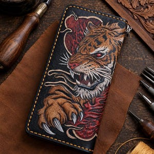Könnte beinhalten: Ein rechteckiges Portemonnaie aus schwarzem Leder mit einem detaillierten, handgefertigten und bemalten Tigermotiv. Der Tiger ist mit gefletschten Zähnen, scharfen Krallen und roten Akzenten dargestellt. Das Portemonnaie hat dicke, sichtbare gelbe Nähte.