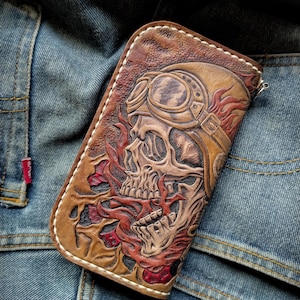 Puede incluir: Una cartera rectangular de cuero marrón con un diseño en relieve de una calavera con gafas de aviador rodeada de llamas rojas. La cartera tiene costuras blancas visibles a lo largo de los bordes y una cremallera plateada en el lateral.