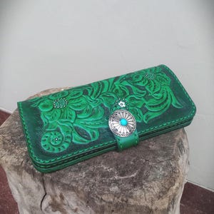 Könnte beinhalten: Grünes Lederportemonnaie mit geprägtem Blumenmuster. Die Geldbörse hat einen silberfarbenen Verschluss mit einem Türkisstein. Das Leder ist in einem leuchtenden Grün gefärbt, und das Design ist mit kleinen silbernen Nieten versehen.