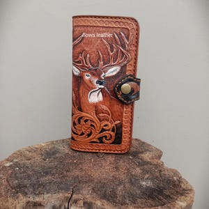 May include: A brown leather wallet with an embossed deer design. The wallet has a floral pattern and a button clasp. The text "flows leather" is visible. The wallet is likely around 10 cm in height.