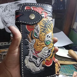 Puede incluir: Cartera de cuero negro con un diseño detallado de cara de tigre en amarillo, naranja y blanco. La cartera tiene un cierre de botón a presión y costuras blancas. Los ojos del tigre son verdes. El diseño está en relieve en la parte delantera.