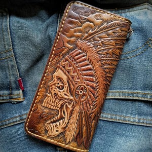 Puede incluir: Una cartera rectangular de cuero marrón con un intrincado diseño en relieve de una calavera con un tocado de plumas. La superficie de cuero muestra tallas detalladas de plumas y patrones geométricos, con costuras visibles a lo largo de los bordes.