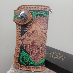 Puede incluir: Cartera de cuero marrón con diseño de león rugiente. La cartera presenta una cabeza de león en relieve detallada, detalles florales verdes y un cierre plateado. La cartera mide aproximadamente 18 cm de largo.