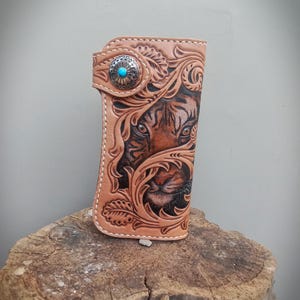 Puede incluir: Una cartera de cuero marrón claro con un tallado detallado de la cara de un tigre. La cartera presenta un cierre de botón turquesa y patrones florales decorativos. Los ojos y la nariz del tigre son visibles, con sombreado intrincado. La cartera se muestra sobre una superficie de madera.
