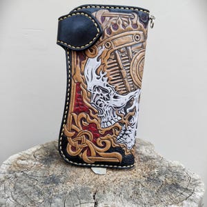 Peut inclure: Un portefeuille en cuir haut et rectangulaire orné de sculptures complexes représentant deux crânes, des motifs de flammes et des entrelacs celtiques. Le cuir est teint en noir, beige, rouge et blanc, avec une fermeture à bouton-pression.