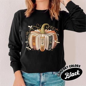May include: Black long-sleeve shirt featuring a pumpkin design made of books. The pumpkin has a green stem and is surrounded by bats and decorative swirls. The text "COMFORT COLORS Black" is in a black oval.