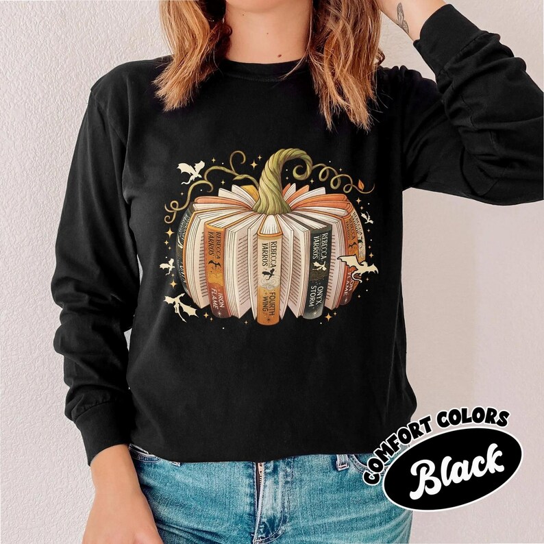May include: Black long-sleeve shirt featuring a pumpkin design made of books. The pumpkin has a green stem and is surrounded by bats and decorative swirls. The text "COMFORT COLORS Black" is in a black oval.