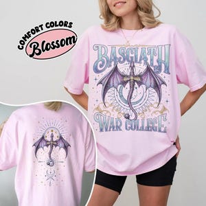 May include: A pale pink t-shirt with a dragon graphic and the words "BASCIATH" and "WAR COLLEGE". The dragon has purple wings and is the central design element. The back of the shirt also features a dragon design.