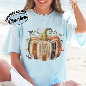 May include: Light blue t-shirt with a pumpkin design made of books. The books have titles like "Iron Flame" and "Onyx Storm." The shirt also features the text "COMFORT COLORS Chambray."