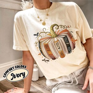 May include: Ivory-colored t-shirt featuring a pumpkin design made of books. The pumpkin has a green stem and is surrounded by bats and stars. The text "Comfort Colors Ivory" is in an oval on the bottom left.