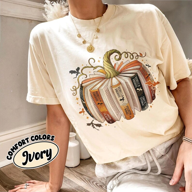May include: Ivory-colored t-shirt featuring a pumpkin design made of books. The pumpkin has a green stem and is surrounded by bats and stars. The text "Comfort Colors Ivory" is in an oval on the bottom left.