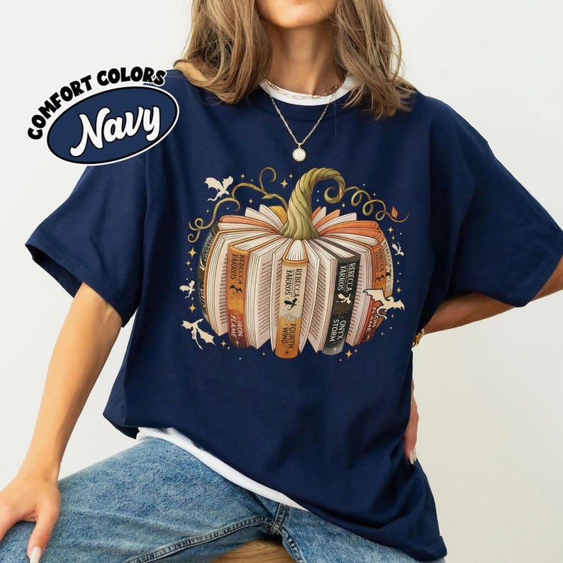 May include: Navy blue t-shirt with a pumpkin design made of books. The books have titles like "Onyx Storm" and "Iron Flame." The shirt also features the text "COMFORT COLORS Navy."