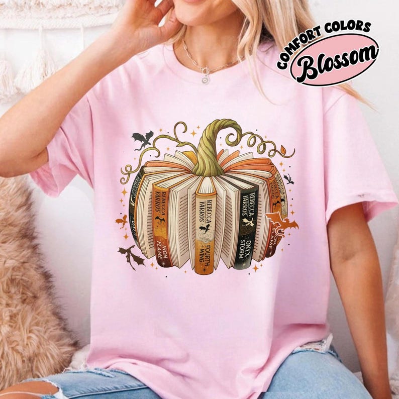 May include: Pink t-shirt featuring a pumpkin design made of books. The books have titles like "Rebecca Yarros" and "Iron Flame." The design includes bats and vines, with the text "COMFORT COLORS Blossom" in a pink oval.
