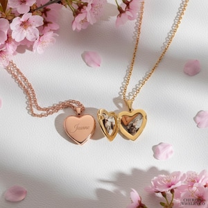 May include: Two heart-shaped lockets on chains, one rose gold and one gold, displayed with pink blossoms. The rose gold locket is engraved with the name "Jeanne." The gold locket is open, revealing a photo inside. The jewelry is on a white surface.
