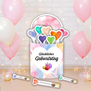 May include: A birthday card with the words "Glücklicher Geburtstag" in black script, featuring colorful watercolor hearts and gift boxes. Pink and white balloons and decorative lights surround the card, creating a festive atmosphere.