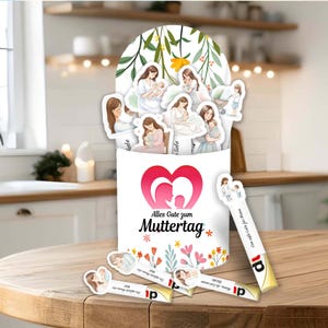 Puede incluir: Una caja blanca con un diseño de corazón y el texto "Alles Gute zum Muttertag". La caja contiene pegatinas de madres y bebés. Varias pegatinas están esparcidas alrededor de la caja. La caja está sobre una superficie de madera.