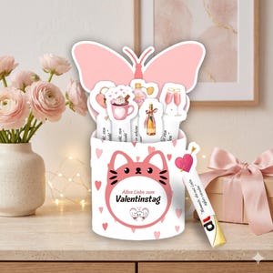 May include: A white container with a pink cat face design and the text "Alles Liebe zum Valentinstag". The container holds several pink and white Valentine's Day themed cutouts, including a butterfly, hearts, and champagne glasses. A gift box with a pink ribbon is in the background.