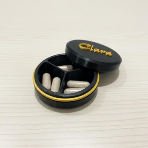 May include: A black and yellow pill organiser with the name "Clara" on the lid. The organiser is divided into three sections and contains several capsules. The lid is open, revealing the contents.