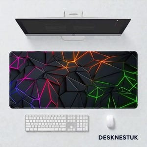 May include: A black desk mat with a geometric pattern and neon lines in pink, orange, and green. A white keyboard, mouse, and computer monitor are also visible. The text "DESKNESTUK" is in the bottom right corner.