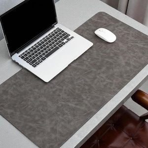 Pode incluir: Um tapete de mesa retangular cinza com uma aparência texturizada semelhante ao couro. Um laptop branco e um mouse sem fio branco estão no tapete. O tapete está em uma mesa cinza claro.