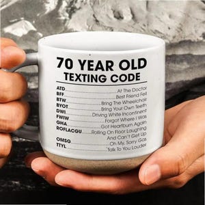 May include: White ceramic mug with a tan base, featuring the text "70 YEAR OLD TEXTING CODE" and a list of acronyms with their meanings. The mug is held by a person.