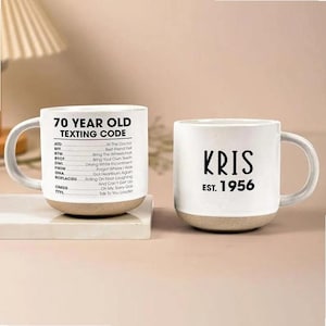 May include: Two ceramic mugs. One mug has the text "70 YEAR OLD TEXTING CODE" with a list of texting acronyms. The other mug has the name "KRIS" and "EST. 1956". Both mugs have a white upper section and a beige base.