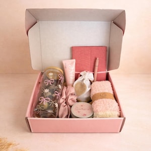 May include: A pink gift box filled with self-care items. Includes a glass with floral and bow designs, a pink notebook, a pen, a tube of lotion, a scrunchie, a small bag, a candle, and a fluffy pink towel.