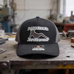 May include: Black and gray trucker hat with the text "POTBELLIED STALLION" in white lettering. The hat features a brown silhouette of a person. The hat is adjustable and has a curved brim.