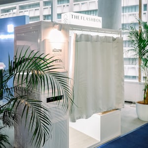 Può includere: Una cabina fotografica bianca con l'etichetta THE FLASHBOX, dotata di una tenda color chiaro, un piccolo faretto e un cartello con il testo allure BEAUTY BOOTH B&W. La cabina ha una struttura rettangolare e un'area d'ingresso aperta per gli utenti.