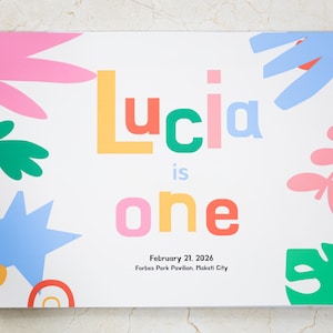 Può includere: Un quaderno a spirale con copertina bianca. La copertina mostra il nome "Lucia" in lettere colorate, insieme alle parole "is one". Il design include forme floreali e geometriche astratte in rosa, verde, blu e arancione.