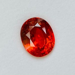 May include: Oval-cut, faceted gemstone with a vibrant orange-red hue. The gem has a polished surface with internal reflections, showcasing its clarity and brilliance. The gemstone is set against a plain white background.