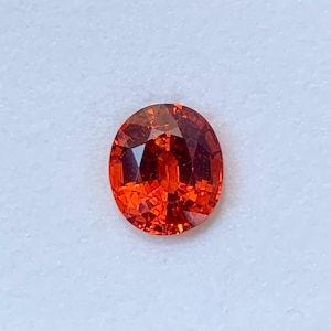 May include: A vivid orange, oval gemstone with a faceted surface. The gem's facets catch the light, producing a sparkling effect. The colour varies from a deep reddish-orange to a bright, fiery hue. The gemstone is set against a plain white background.
