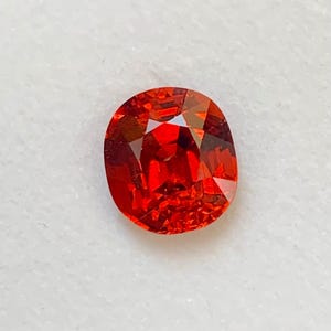 May include: A vivid, oval-shaped, red gemstone with a faceted surface. The gem exhibits a rich, fiery colour and is set against a white background. The gemstone's facets catch the light, creating a sparkling effect.