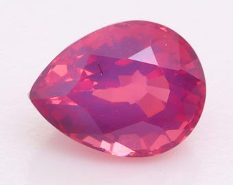 Puede incluir: Una piedra preciosa en forma de pera con un tono rosa rojizo vibrante y saturado. La piedra presenta una superficie facetada que crea múltiples reflejos geométricos y refracciones de luz en su cuerpo pulido y translúcido, mostrando una silueta clara en forma de lágrima.
