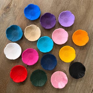 May include: A collection of fifteen small, circular silicone bowls shaped like flowers, each featuring a textured center and smooth petals. The bowls are presented in a variety of solid colors including blue, purple, lavender, beige, pink, orange, white, magenta, cyan, yellow, navy, red, dark green, light pink, and black.