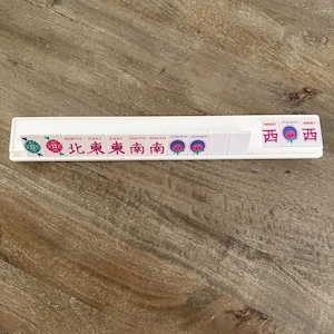 May include: A white plastic Mahjong tile rack holding a sequence of tiles. The tiles display various symbols, including red and green floral designs, the words NORTH, EAST, SOUTH, JOKER, and WEST, along with Chinese characters for compass directions.