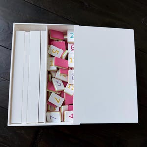 May include: A white rectangular storage box with a sliding lid, containing a set of small rectangular tiles. The tiles feature pink backs and white fronts printed with colorful numbers in various fonts, alongside several plain white rectangular dividers.