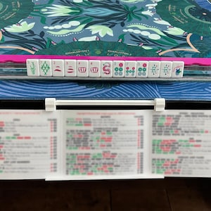 May include: A Mahjong rack holding a row of white tiles with green, red, and blue symbols, including numbers and characters. The rack is positioned on a blue patterned mat with a white card holder displaying a blurry printed score card.