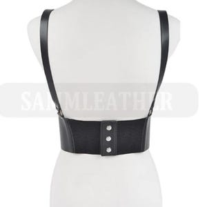 Puede incluir: Arnés de cuero negro con tirantes y cintura elástica. El arnés presenta tres botones plateados en la parte delantera. El nombre de la marca "SAMMLEATHER" es visible en el fondo.