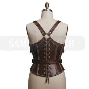 May include: Brown leather corset with adjustable straps and a laced front. The corset features a belt around the waist and decorative stitching. The straps cross in the back and attach to the corset with metal rings. The corset is displayed on a mannequin.