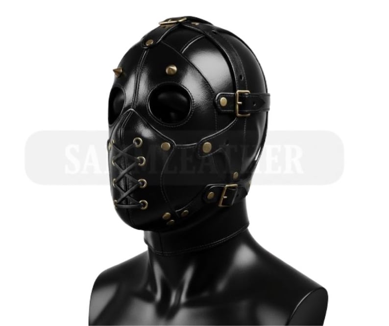 May include: Black leather mask with eye and mouth openings, secured with straps and buckles. The mask features metal studs and rivets, and a lace-up closure on the lower face. The mask is designed to cover the entire head and neck.