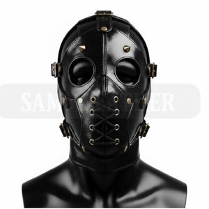 May include: A black leather mask with eye and mouth openings, secured with straps and rivets. The mask features a lace-up design on the lower face and is designed to cover the entire head. The mask is displayed on a black mannequin head.