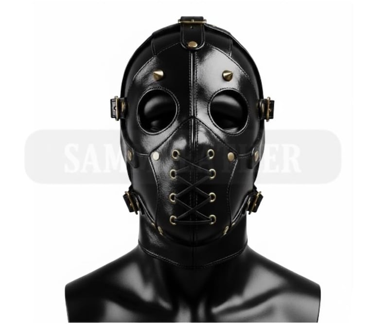 May include: A black leather mask with eye and mouth openings, secured with straps and rivets. The mask features a lace-up design on the lower face and is designed to cover the entire head. The mask is displayed on a black mannequin head.