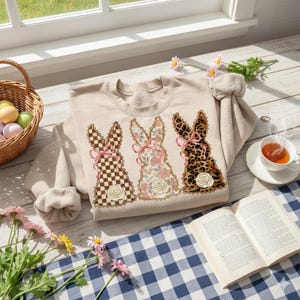 May include: A beige sweatshirt featuring three bunny designs with different patterns: checkered, floral, and leopard print. Each bunny has a pink bow and a rose detail. The sweatshirt is displayed on a blue and white checkered cloth, with a basket of Easter eggs and a cup of tea nearby.