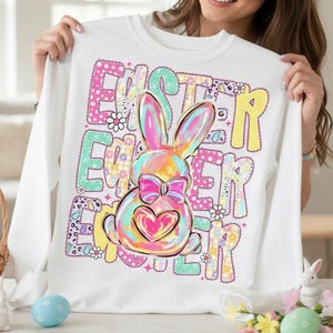 May include: White long-sleeved sweatshirt featuring a colourful Easter bunny design. The word "EASTER" is printed in large, multi-coloured letters. The bunny has a bow and a heart on its belly, with floral accents.